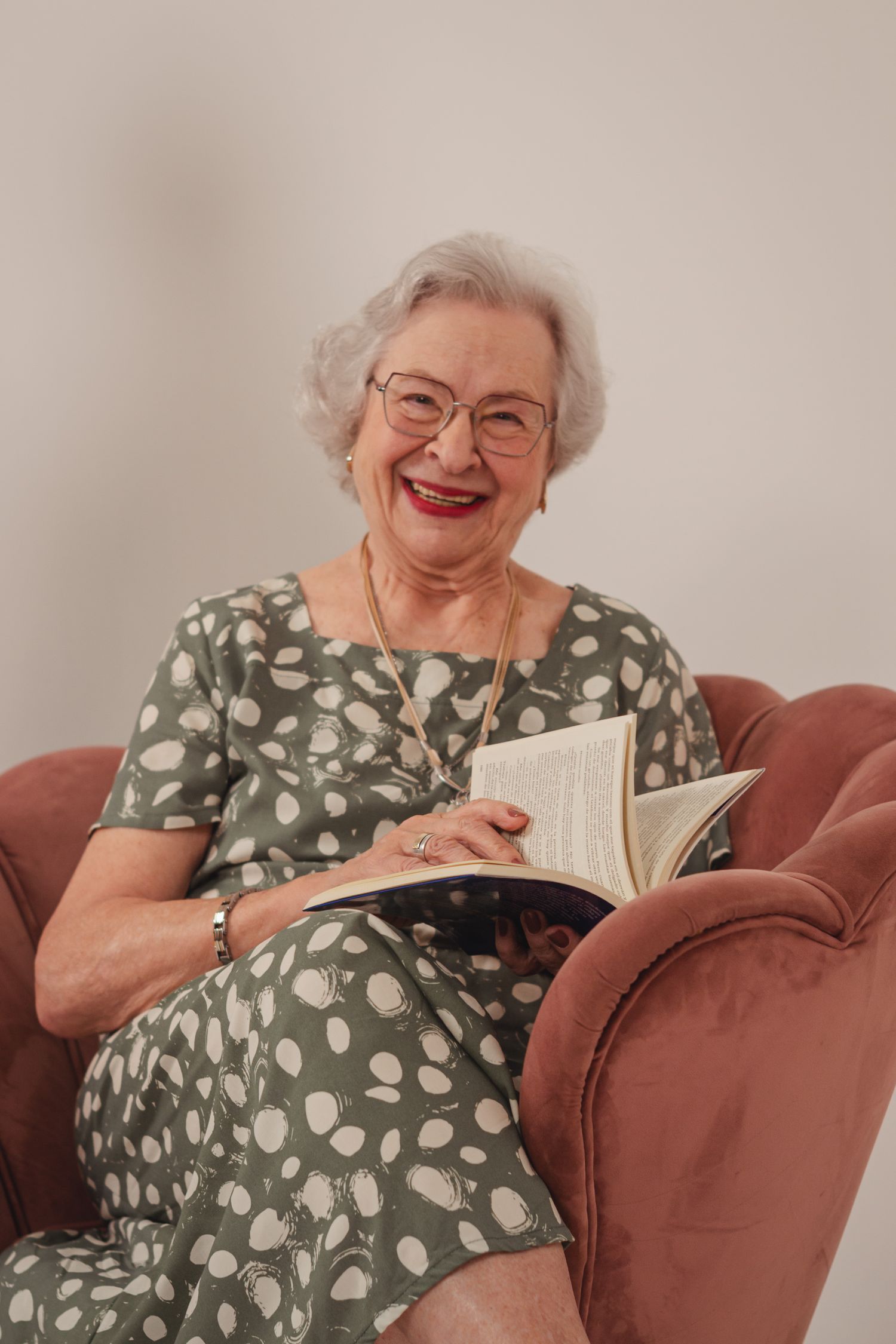 An older woman in a floral dress smiles in an armchair with an open book on her lap