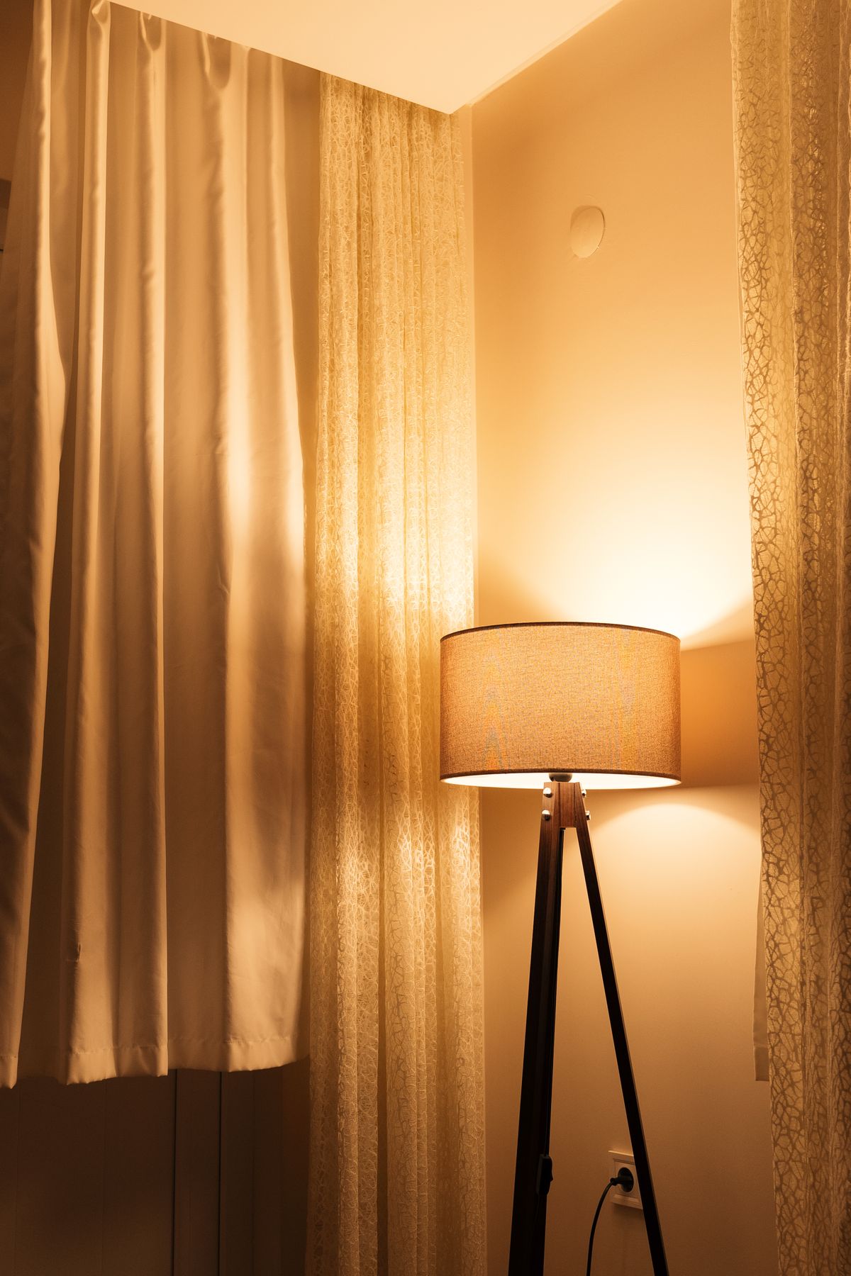 A floor lamp glowing warm beside drawn curtains in a room just after dusk